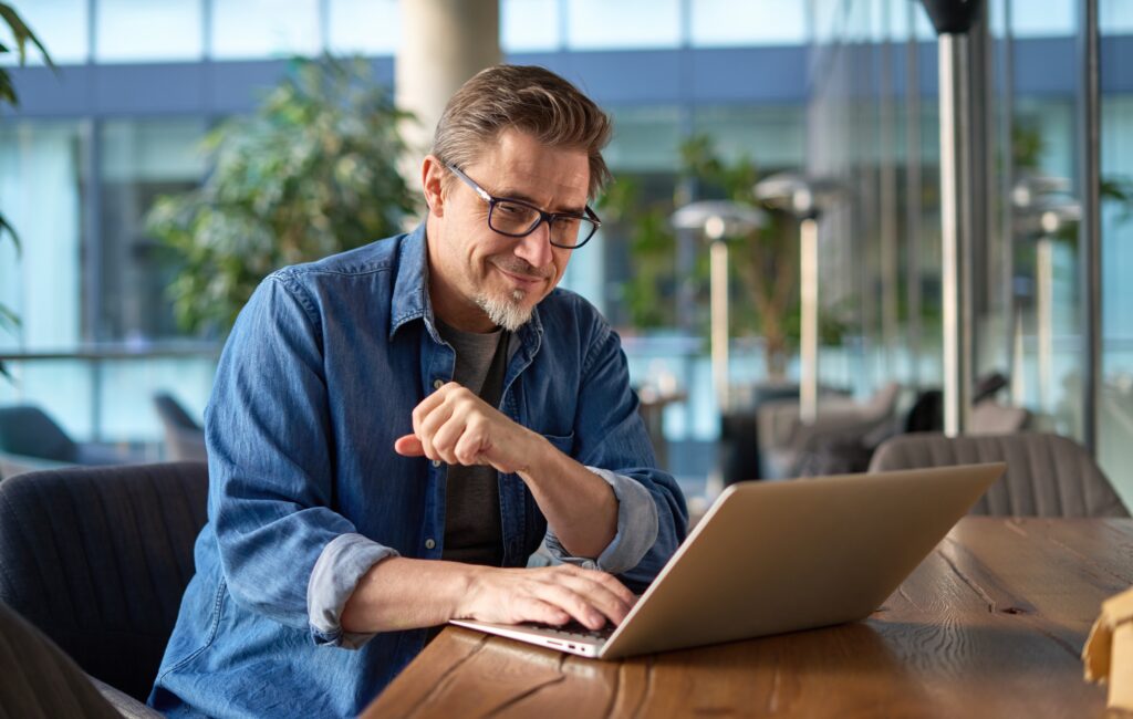 Happy middle age businessman using laptop computer in office. Older casual man working online on terrace of cafe, smiling.